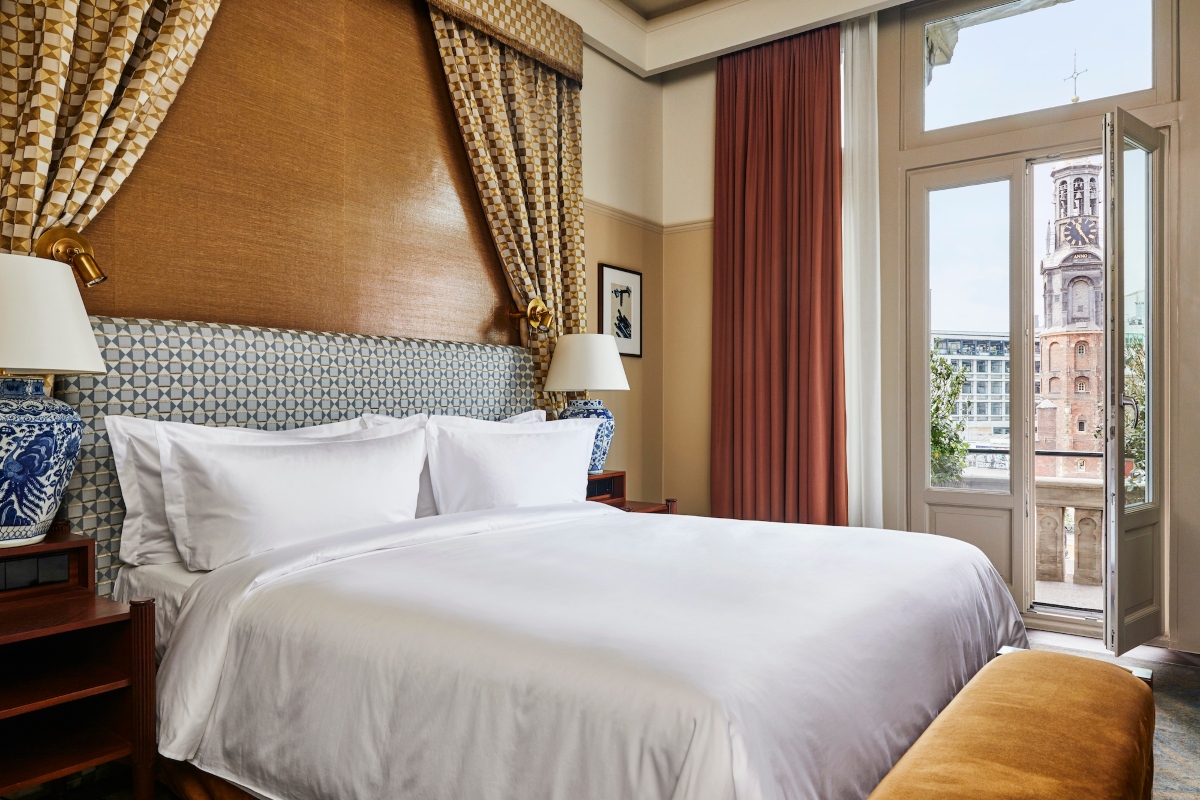 A hotel room features a bed with crisp white linens, a blue-and-white geometric upholstered headboard framed by gold patterned drapes, and blue-and-white porcelain lamps on wooden nightstands. Tall French doors with rust-colored curtains open onto a small balcony overlooking a brick clock tower.