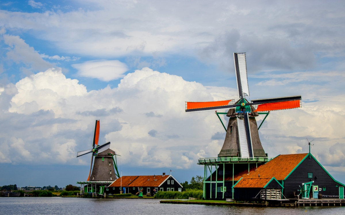 Two traditional windmills with thatched-style gray bodies, green trim, and white and orange sails stand along the edge of a calm river, each beside a dark wooden building with an orange tile roof. Towering white cumulus clouds fill a blue sky above a flat green shoreline in the distance.