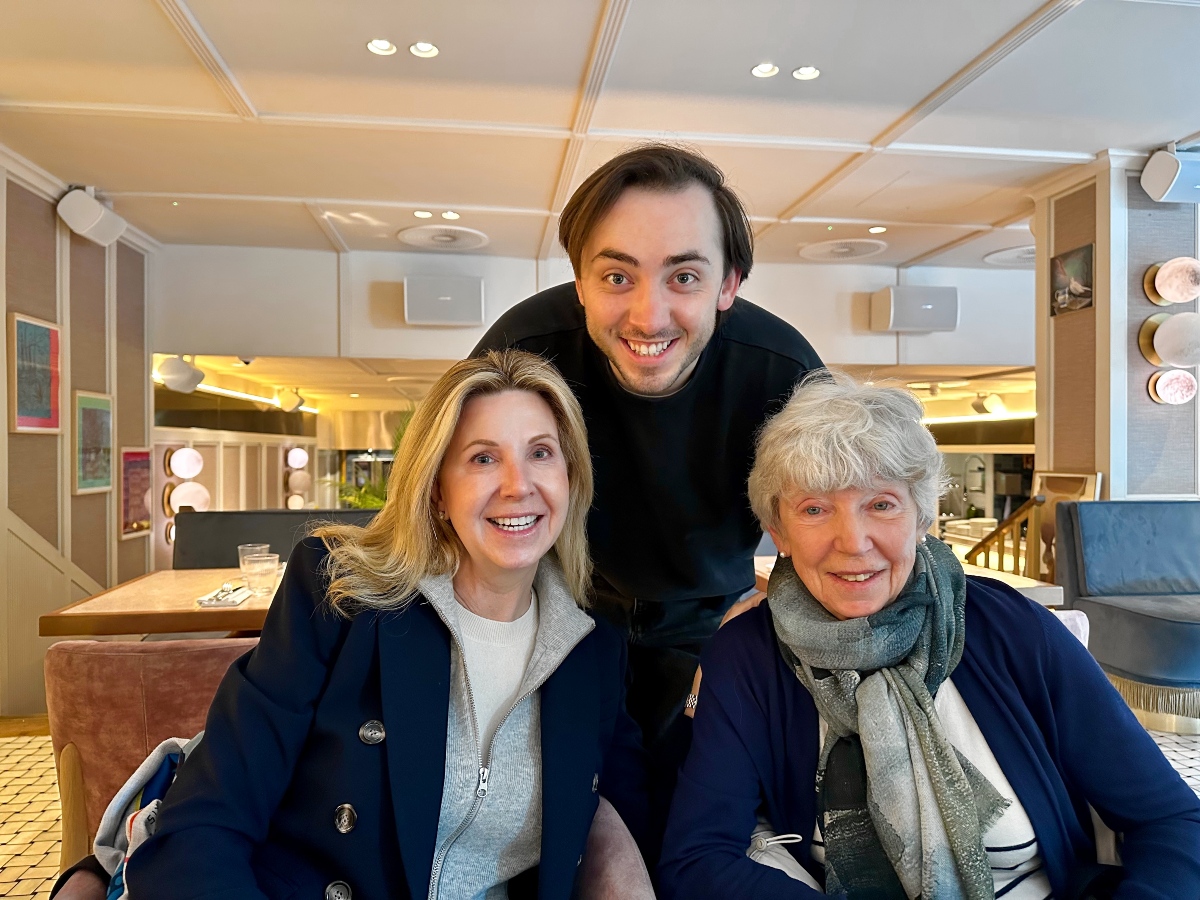Two women sit at a table in a bright, modern restaurant, smiling at the camera, while a young man with dark hair in a black sweater leans in between them from behind with a wide grin. The woman on the left has shoulder-length blonde hair and wears a navy coat over a gray zip-up sweater, and the woman on the right has short silver hair and wears a navy cardigan with a gray-green scarf, with pastel wall art, round wall lights, and blue velvet seating in the background.