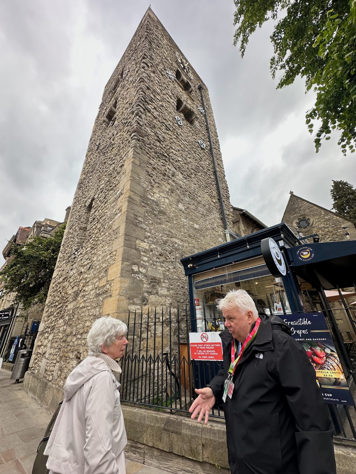 A white-haired man in a black jacket and lanyard gestures as he speaks with a woman with short silver hair in a light gray hooded coat, standing on the pavement beside iron railings at the base of a tall, rough stone church tower with a clock face near the top. A navy crêpe kiosk with a sign reading "Irresistible Crepes" sits beside the tower under an overcast sky, with tree branches framing the upper right of the scene.