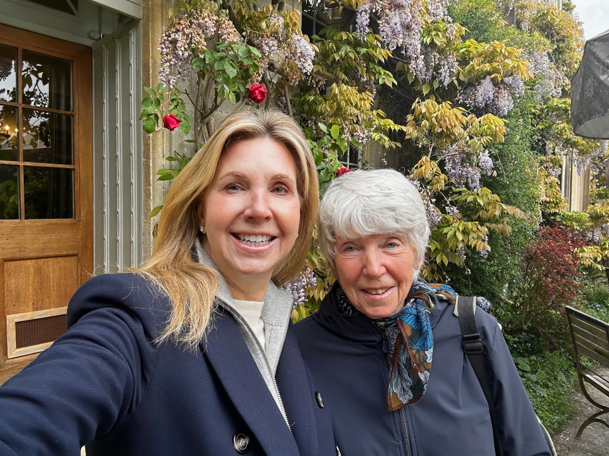 Two women smile in a close-up selfie outside a honey-colored stone building, the woman on the left with shoulder-length blonde hair in a navy coat over a gray zip-up, and the woman on the right with short silver hair in a navy jacket and a blue-and-rust patterned scarf. Behind them, lavender wisteria and red climbing roses cascade over the wall beside a glass-paneled wooden door, with a wooden bench and garden greenery to the right.
