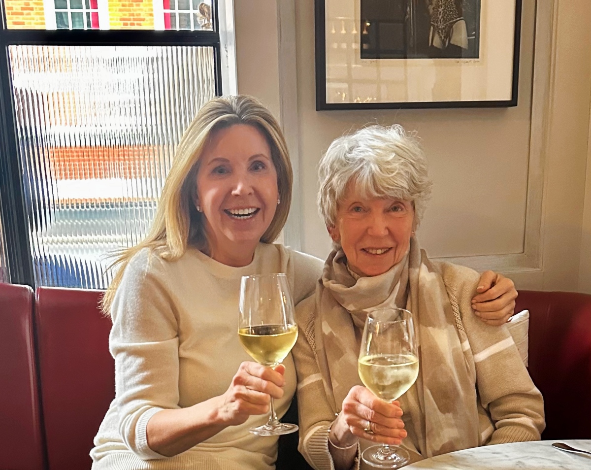 Two women sit side by side on a red leather banquette in a restaurant, smiling and raising glasses of white wine toward the camera, with one woman's arm around the other's shoulders. The woman on the left has shoulder-length blonde hair and wears a cream sweater, while the woman on the right has short silver hair and wears a beige patterned scarf, with a ribbed glass window and a framed black-and-white photograph behind them.