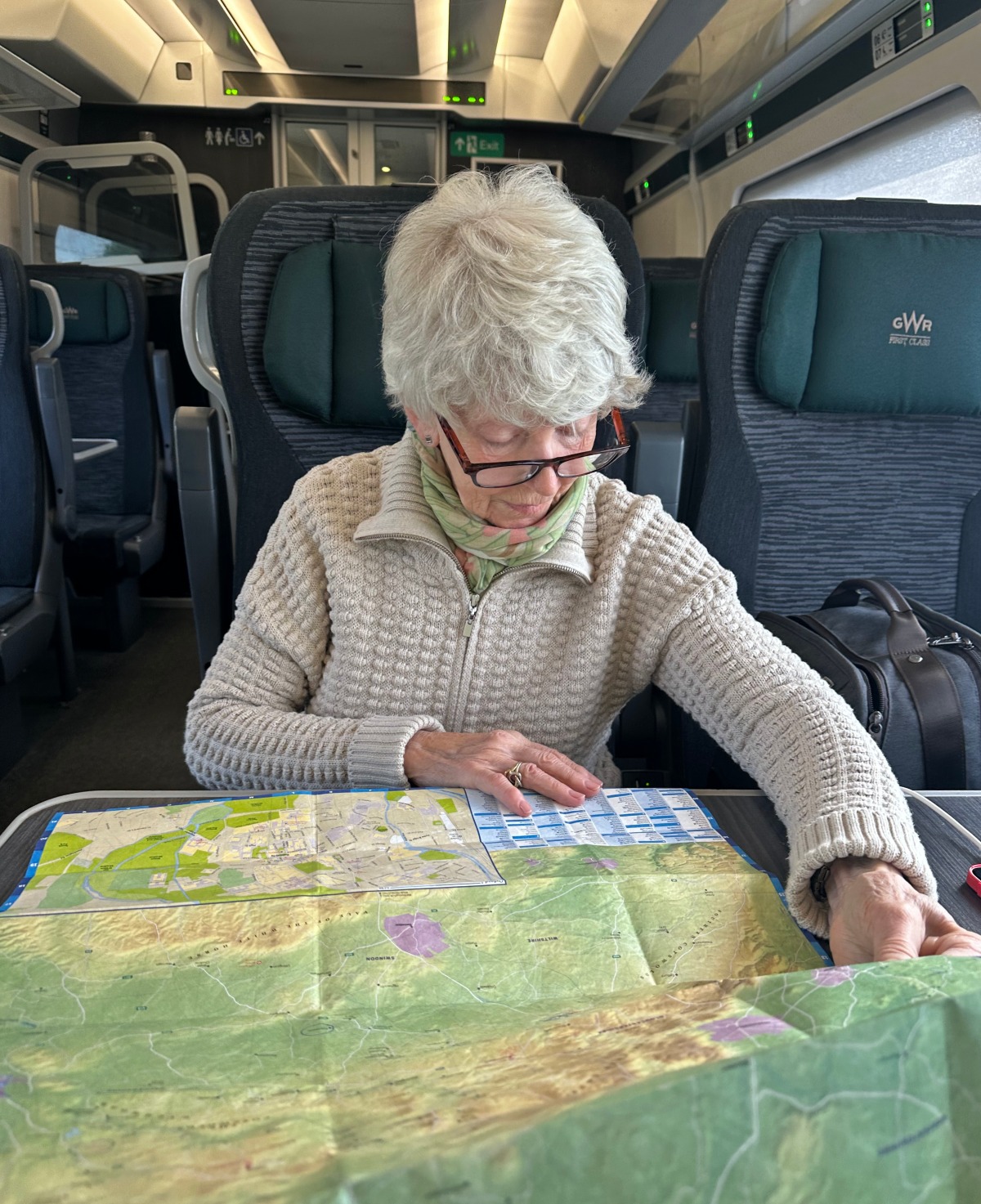 A woman with short silver hair and tortoiseshell glasses studies a large unfolded map spread across a train table, wearing a cream textured zip-up sweater and a green-and-pink patterned scarf. She sits in a teal-and-navy upholstered seat with "GWR First Class" embroidered on the headrest beside her, with a dark bag on the adjacent seat and the carriage's empty rows and overhead luggage racks behind her.