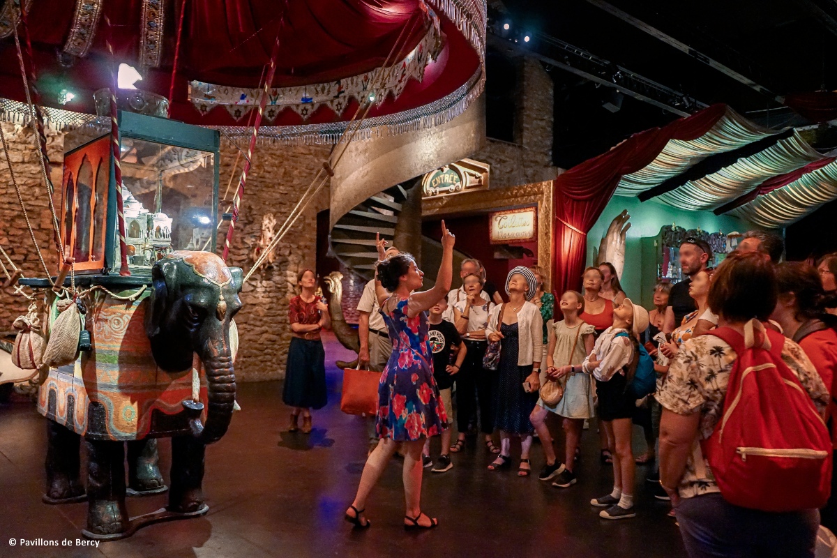 A guide in a colorful floral dress points upward as a group of adults and children gazes toward the ceiling in a dimly lit exhibit hall with stone walls and draped red and gold canopies. At left, a carved elephant with an ornate saddle blanket carries a lantern-like glass display case, and a spiral staircase and an "Entrée" sign are visible in the background.