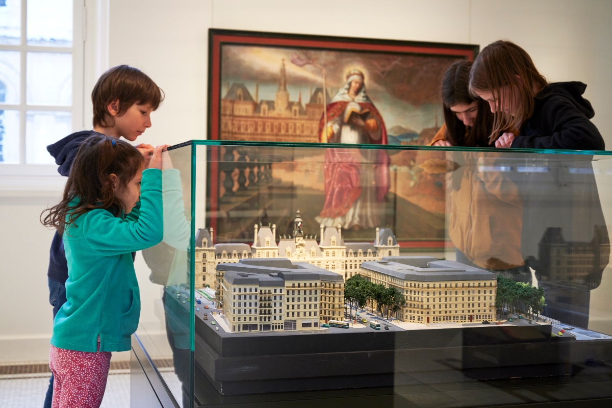 Four children lean over and peer into a glass display case holding a detailed scale model of Parisian buildings, including an ornate, turreted palace-like structure, cream-colored apartment blocks, trees, and tiny buses and cars. Behind them hangs a large framed painting of a haloed woman in red robes holding an open book, with a grand building and a river in the background.