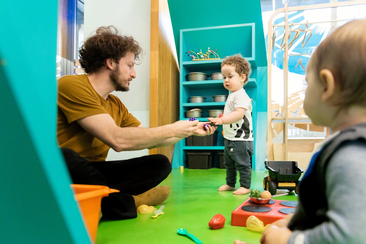 A bearded man in a mustard-colored T-shirt sits cross-legged on a bright green floor, handing a small purple toy to a barefoot toddler with curly hair who reaches out to take it. Play food, a toy stove, a dump truck, and scattered utensils surround them, with turquoise shelves of metal bowls in the background and a second baby watching in the foreground.