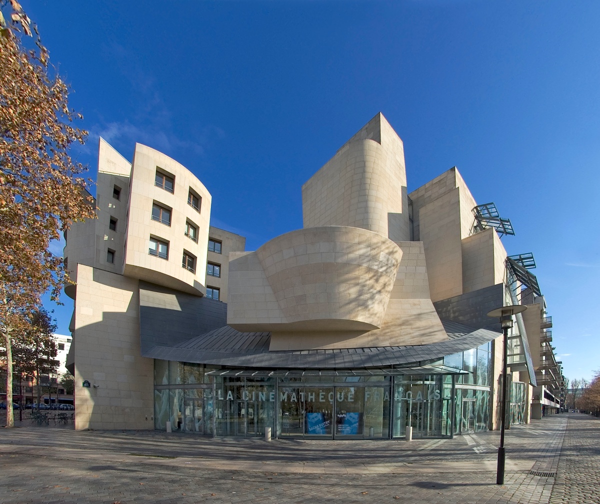 A sculptural, cream-colored stone building with curving walls, jutting angular towers, and a sweeping zinc canopy rises against a bright blue sky. "La Cinémathèque Française" is lettered across the glass entrance at ground level, with a wide cobblestone plaza, a lamppost, and autumn trees in the foreground.