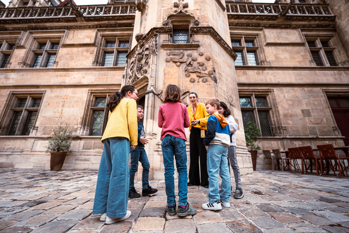 A museum guide in a yellow top speaks to a small group of children gathered on the cobblestone courtyard of a Gothic stone mansion. Behind them rises an ornately carved stair tower decorated with scallop-shell motifs, flanked by tall leaded-glass windows and a stone balustrade along the roofline.