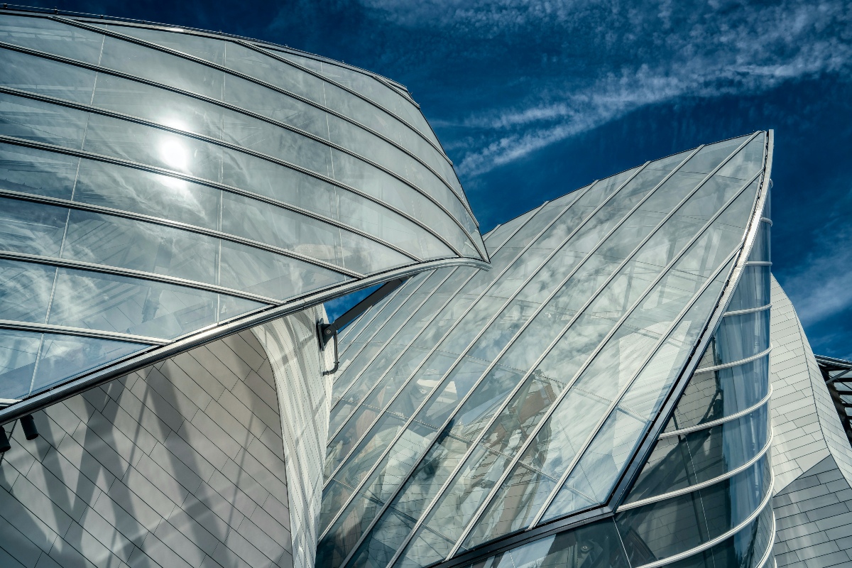 A close-up, upward view of the Fondation Louis Vuitton's architecture shows sweeping, curved glass panels shaped like billowing sails, supported by slender metal ribs against a deep blue sky streaked with wispy clouds. Beneath the glass, pale gray tiled surfaces curve and angle in multiple directions, with the sun glinting off the upper left panel.