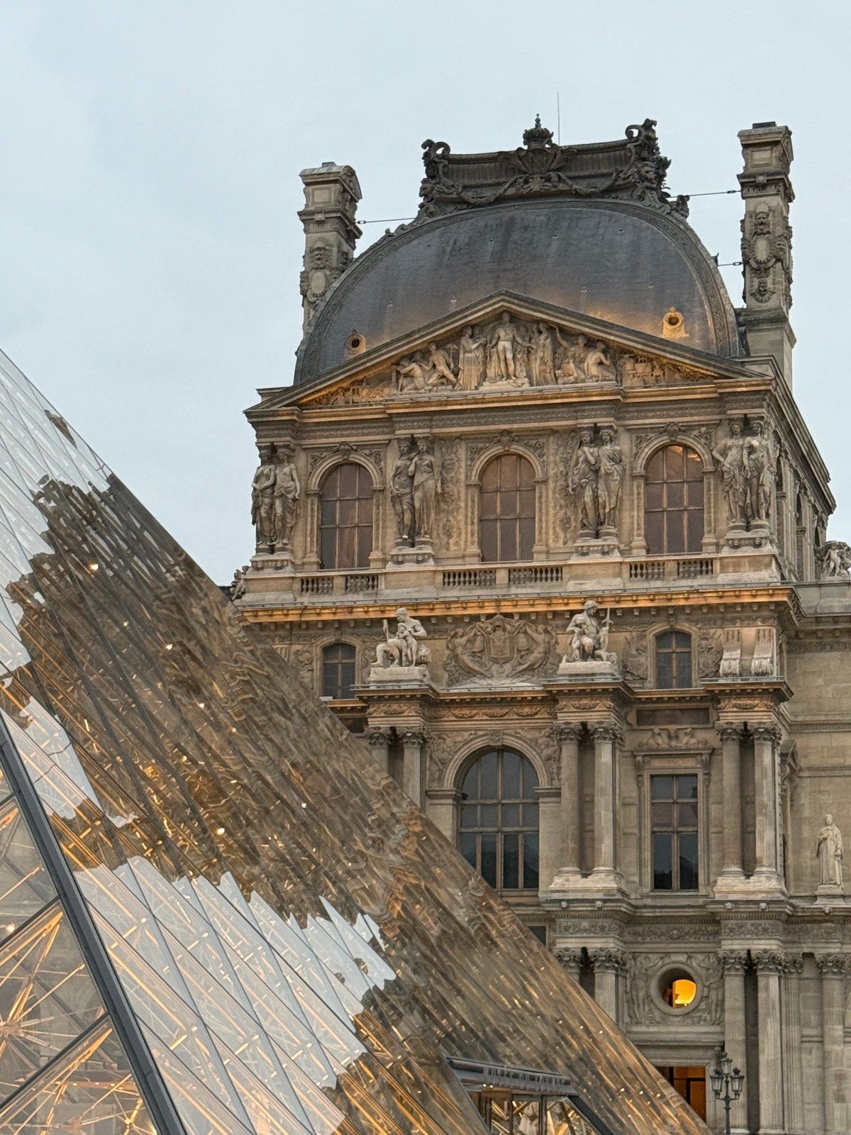 The slanted glass panes of the Louvre Pyramid, lit warmly from within, reflect the ornate stone facade of the Louvre Palace behind them at dusk. The palace pavilion features a dark domed roof, a sculpted pediment, arched windows, and pairs of carved statues standing between the columns.
