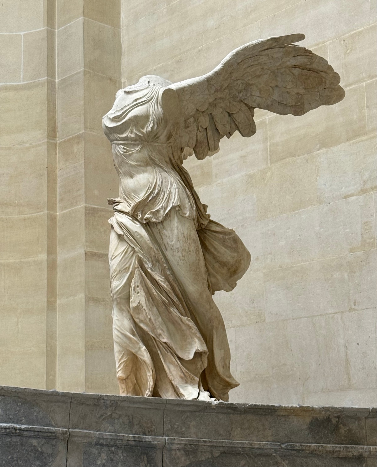 The Winged Victory of Samothrace, a headless and armless ancient marble statue of a winged female figure, stands on a gray stone ship's-prow base against a pale limestone wall. Her outstretched wings sweep back behind her, and her thin, finely carved robes cling to her body and ripple as if blown by strong wind.