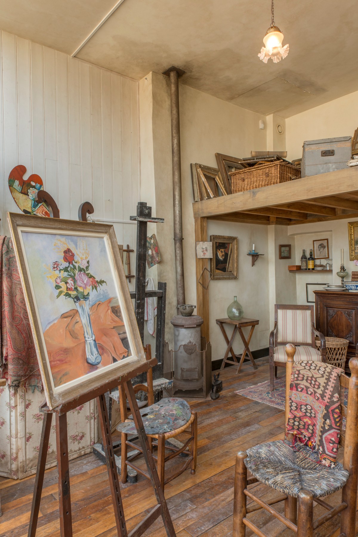 A sunlit artist's studio with worn wooden floors holds a floral still-life painting on a wooden easel, a paint-spattered palette resting on a rush-seat chair, and a tall cylindrical wood stove with a metal pipe rising to the ceiling. A wooden loft stacked with empty picture frames, a wicker basket, and an old trunk sits above a cozy corner with a striped armchair, a carved wooden cabinet, and framed portraits on the walls.