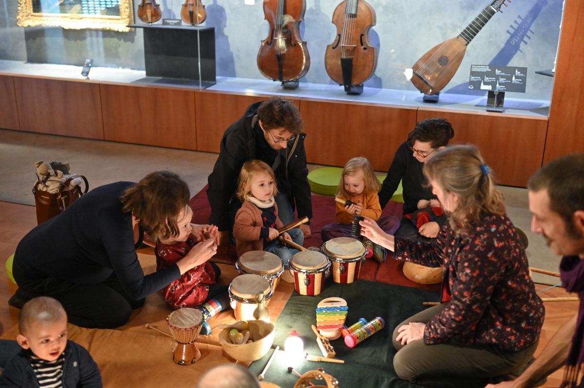 Toddlers and adults sit together on floor mats, playing small hand drums, shakers, a toy xylophone, and other percussion instruments during a group music activity. Behind them, a glass display case holds antique stringed instruments, including cellos, viols, and a lute.