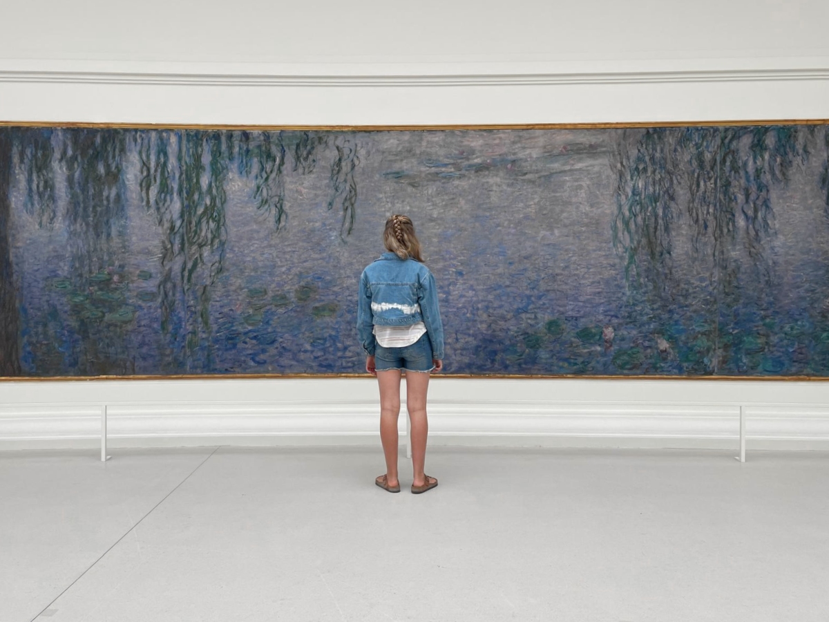 A young girl in a denim jacket, denim shorts, and sandals stands with her back to the camera, facing an enormous, curved Claude Monet Water Lilies panel in a bright white gallery. The painting stretches wider than the frame, showing a hazy blue-violet pond dotted with green lily pads and framed by trailing willow branches on either side.