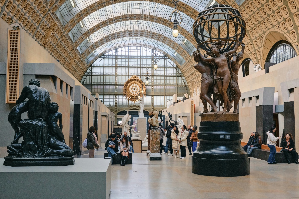 Visitors stroll and sit along the sculpture-lined central nave of the Musée d'Orsay, beneath a soaring arched glass roof framed by gilded, coffered stonework. A bronze group of four figures holding up an armillary globe stands in the right foreground, a dark sculpture of a seated man surrounded by children sits at left, and an ornate gold clock hangs against the tall glass wall at the far end of the hall.