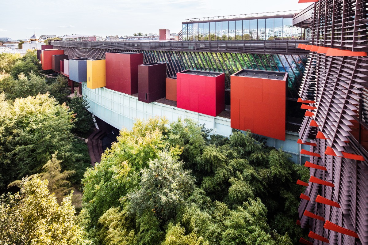 A long, elevated museum building with a glass facade is dotted with protruding boxes painted in bold shades of red, orange, yellow, burgundy, and blue, while angled wooden louvers accented with red panels line the right side. Lush, dense trees and greenery fill the garden beneath the building, and Paris rooftops, including golden onion domes, appear in the distance.