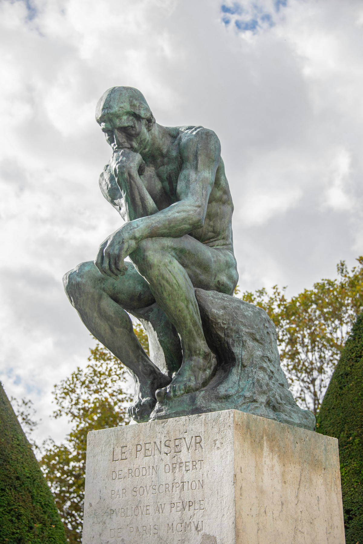 Auguste Rodin's bronze sculpture The Thinker, a muscular nude man seated on a rock with his chin resting on his hand, sits atop a weathered stone pedestal against a cloudy sky. The green-patinated figure is framed by clipped topiary hedges and autumn-tinged trees, and the pedestal is engraved in French with "Le Penseur de Rodin" and a dedication dated MCMVI.