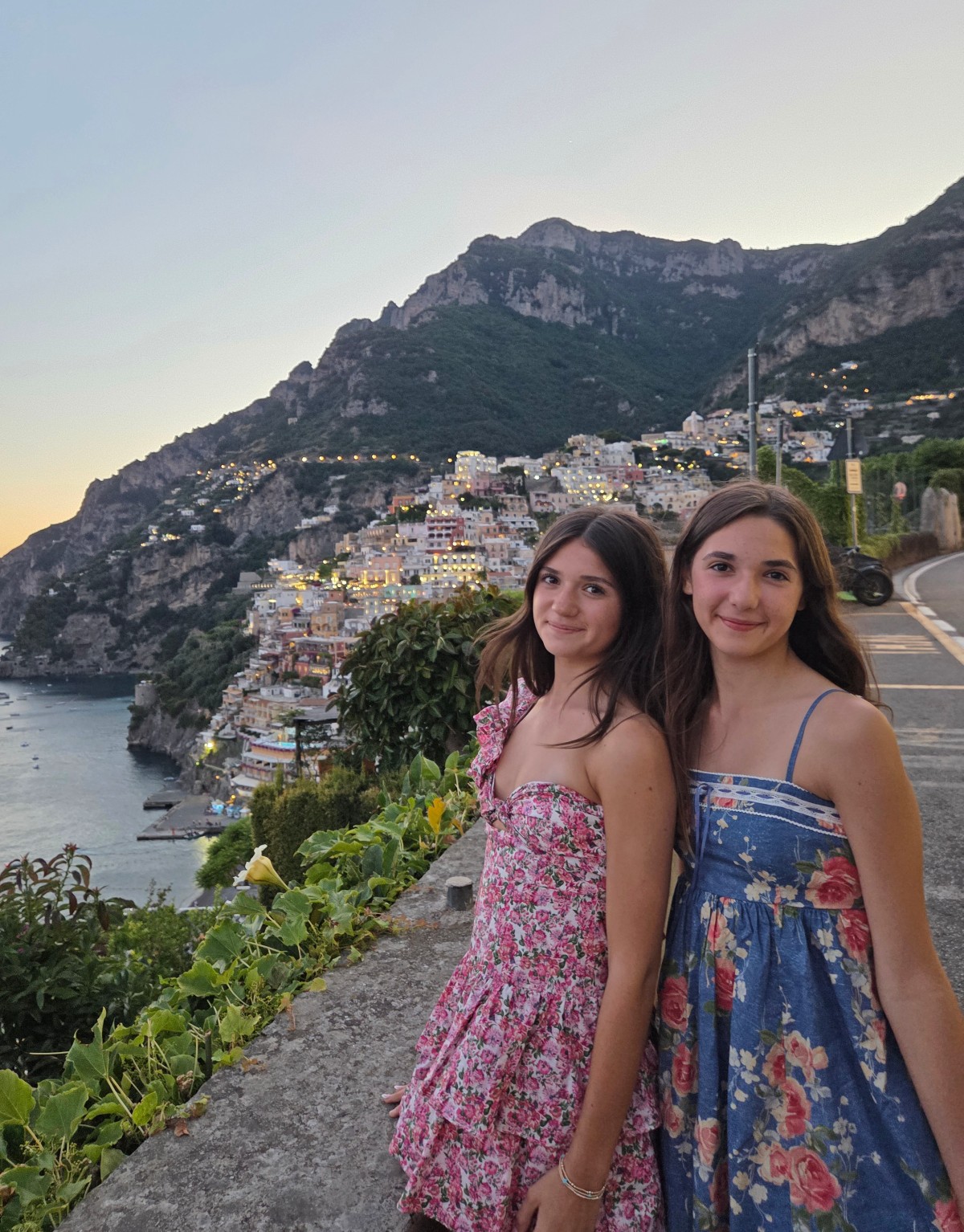 Two girls in floral sundresses, one pink and one blue, stand side by side on a stone overlook wall above Positano at dusk. Behind them, the town's pastel buildings climb a steep green hillside with windows and streetlights glowing, and the harbor and sea are visible below.