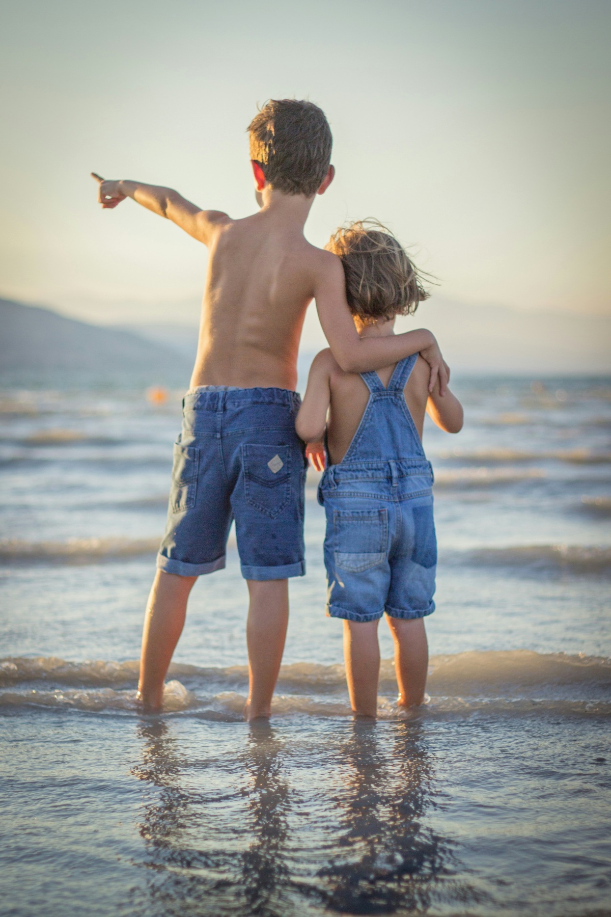 Two young children stand ankle-deep in shallow water at the edge of a beach, seen from behind, with the older one's arm around the younger one's shoulders and pointing out toward the sea. Both wear denim — shorts on the older child, overalls on the younger — and the water reflects warm light from a low sun, with a faint hillside on the horizon.