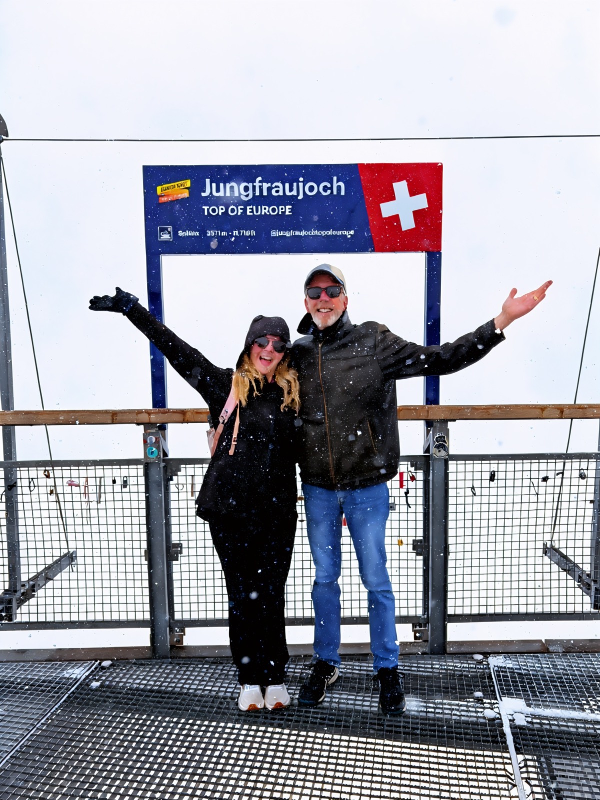 Two adults in winter jackets stand on a metal observation deck with their arms outstretched, smiling, as snow falls around them. Behind them is a blue and red sign reading "Jungfraujoch — Top of Europe" with the Swiss flag, marking an elevation of 3,571 metres, and the view beyond the railing is completely white with cloud.