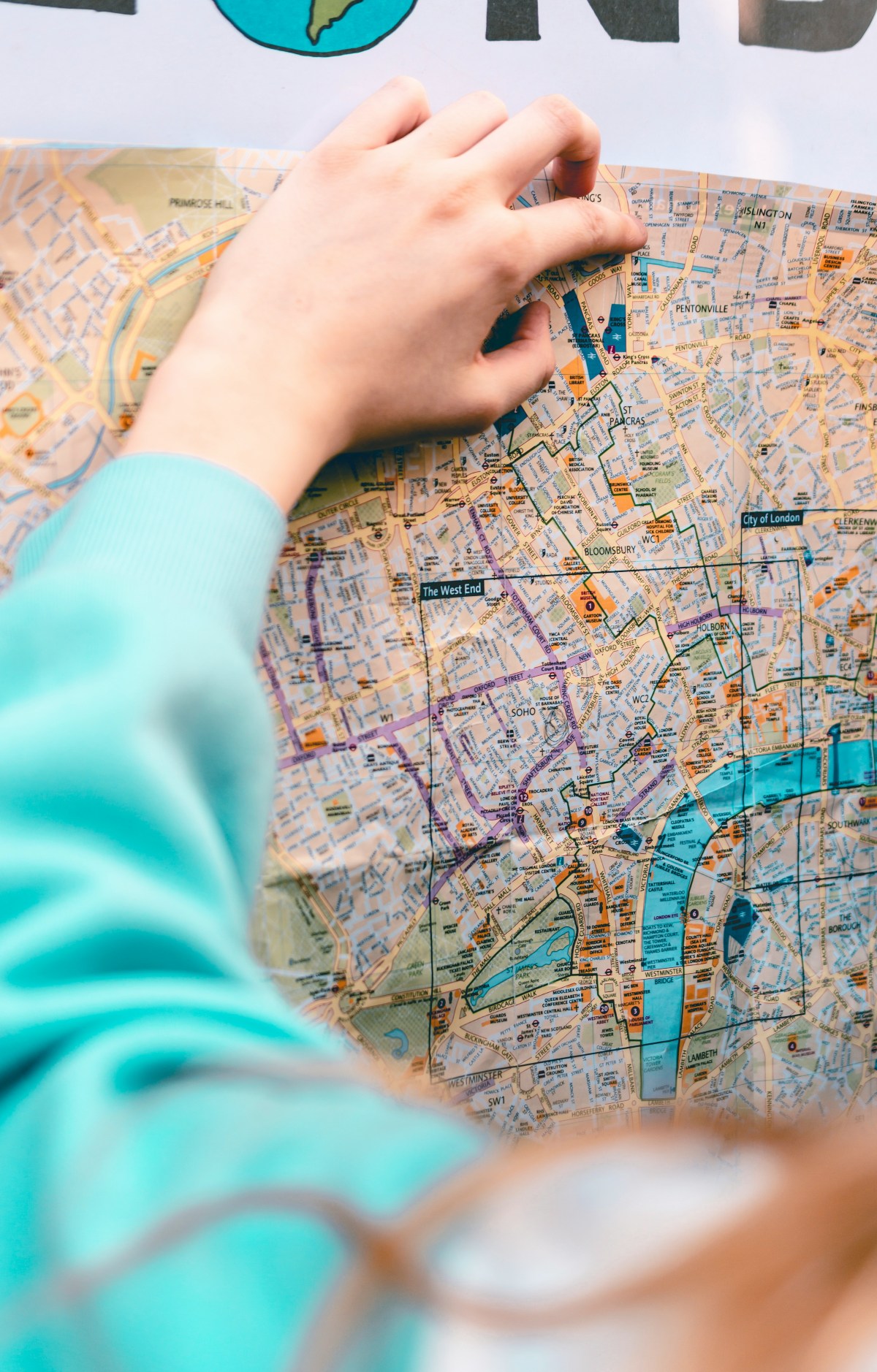 A child in a light blue sweatshirt leans over a folded paper street map of central London, one hand resting on the page near King's Cross. Neighborhood labels including Soho, Bloomsbury, Holborn, and Westminster are visible across the map, along with the Thames marked in blue.