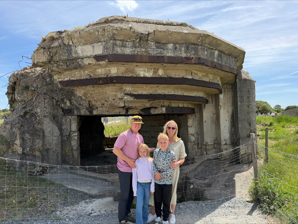 A family of four — two adults and two children — stands together on a gravel path in front of a large concrete German gun emplacement in Normandy, France. The weathered bunker shows exposed rebar, cracked walls, and rusted steel beams above its open firing bay, with grass growing over the roof and a low wire fence running along the front.