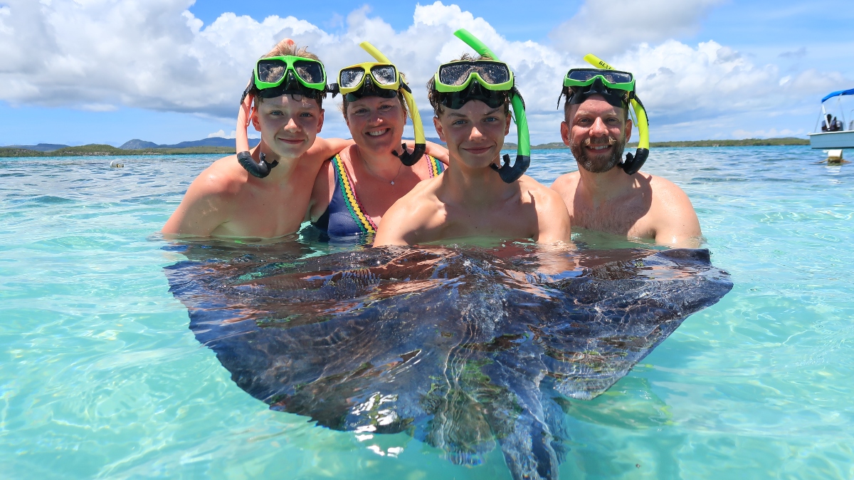 A family of four in snorkel masks stands chest-deep in clear turquoise water, smiling at the camera, with a large stingray floating just below the surface directly in front of them. A low green shoreline and a boat are visible in the distance under a partly cloudy sky.