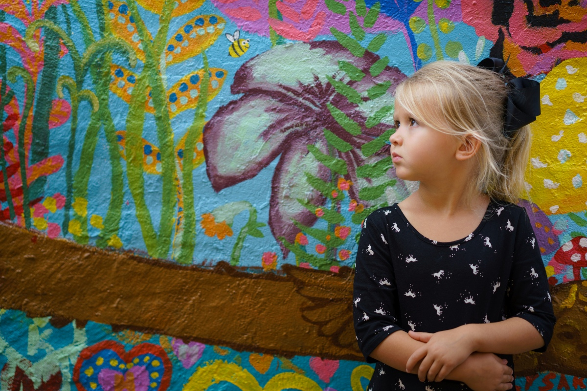 A young girl with blonde hair and a black hair bow stands with her arms crossed in front of a brightly painted mural, looking off to one side. The mural behind her shows a large purple and green flower, a bee, a tree branch, and stylized plants and hearts against a blue background.
