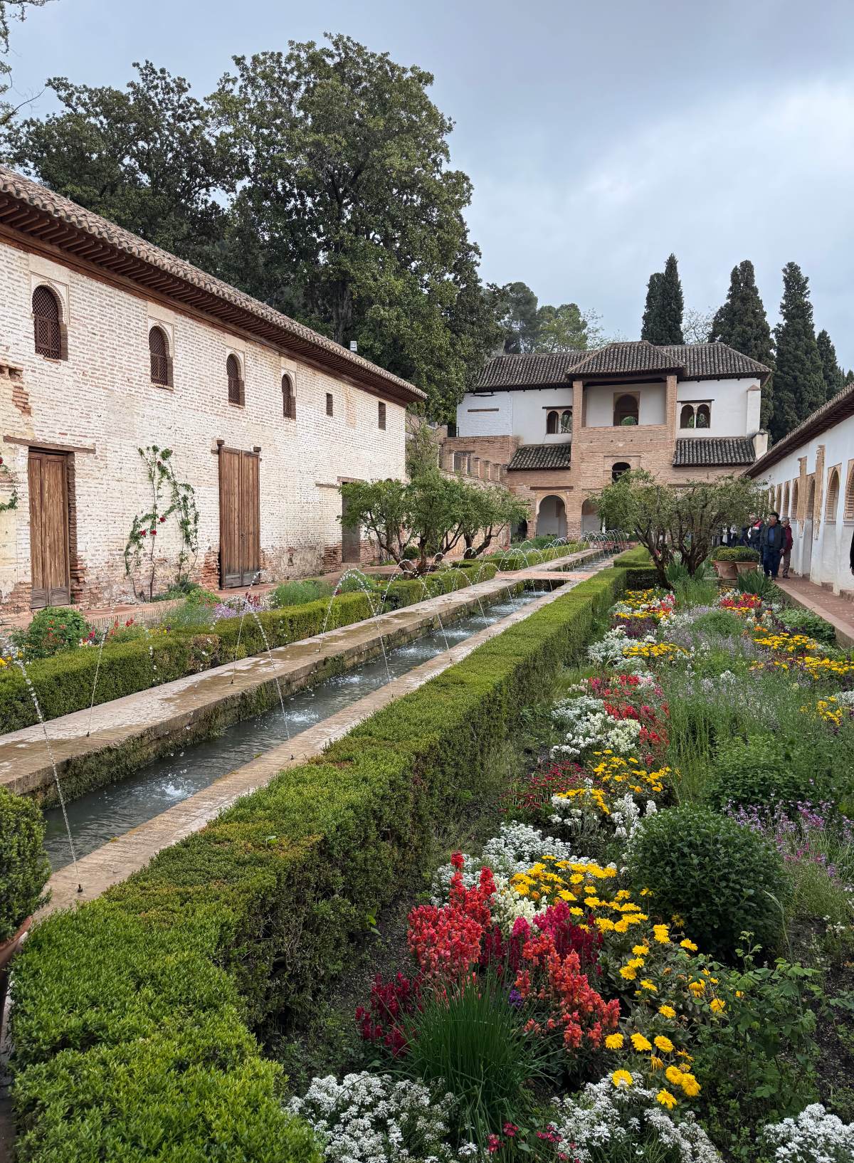 A long, narrow water channel lined with arching fountain jets runs through the Patio de la Acequia in the Generalife gardens at the Alhambra in Granada, Spain, framed by trimmed hedges and beds of red, yellow, and white flowers. Whitewashed and brick buildings with arched windows and tiled roofs line the courtyard, with tall cypress trees and a cloudy sky behind them.