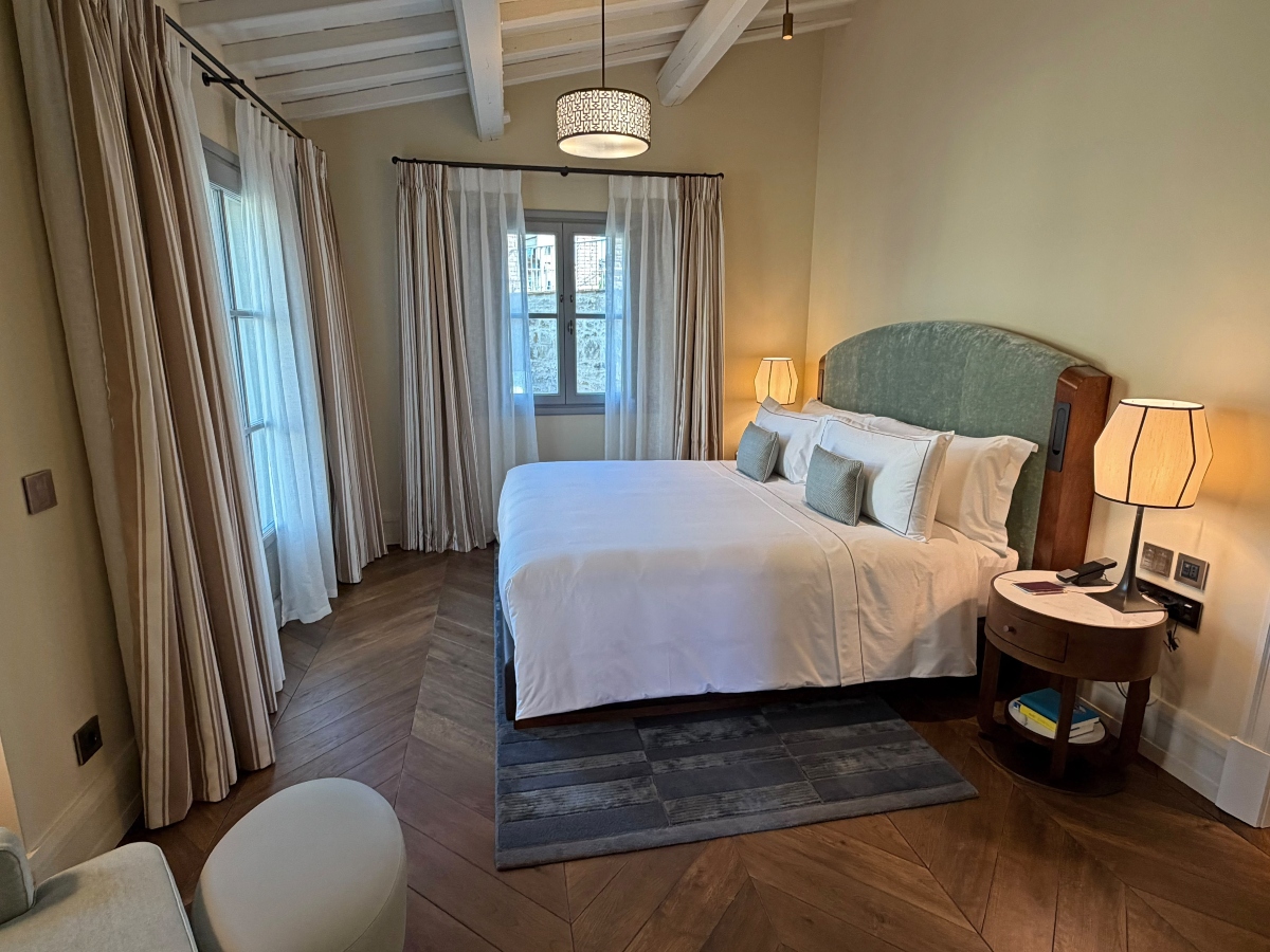 A hotel bedroom features a king bed with crisp white linens and a sage green velvet headboard, flanked by a round wooden nightstand and glowing table lamps. White-beamed ceilings, herringbone wood floors, a gray patterned rug, and tall windows framed by striped and sheer curtains complete the room.