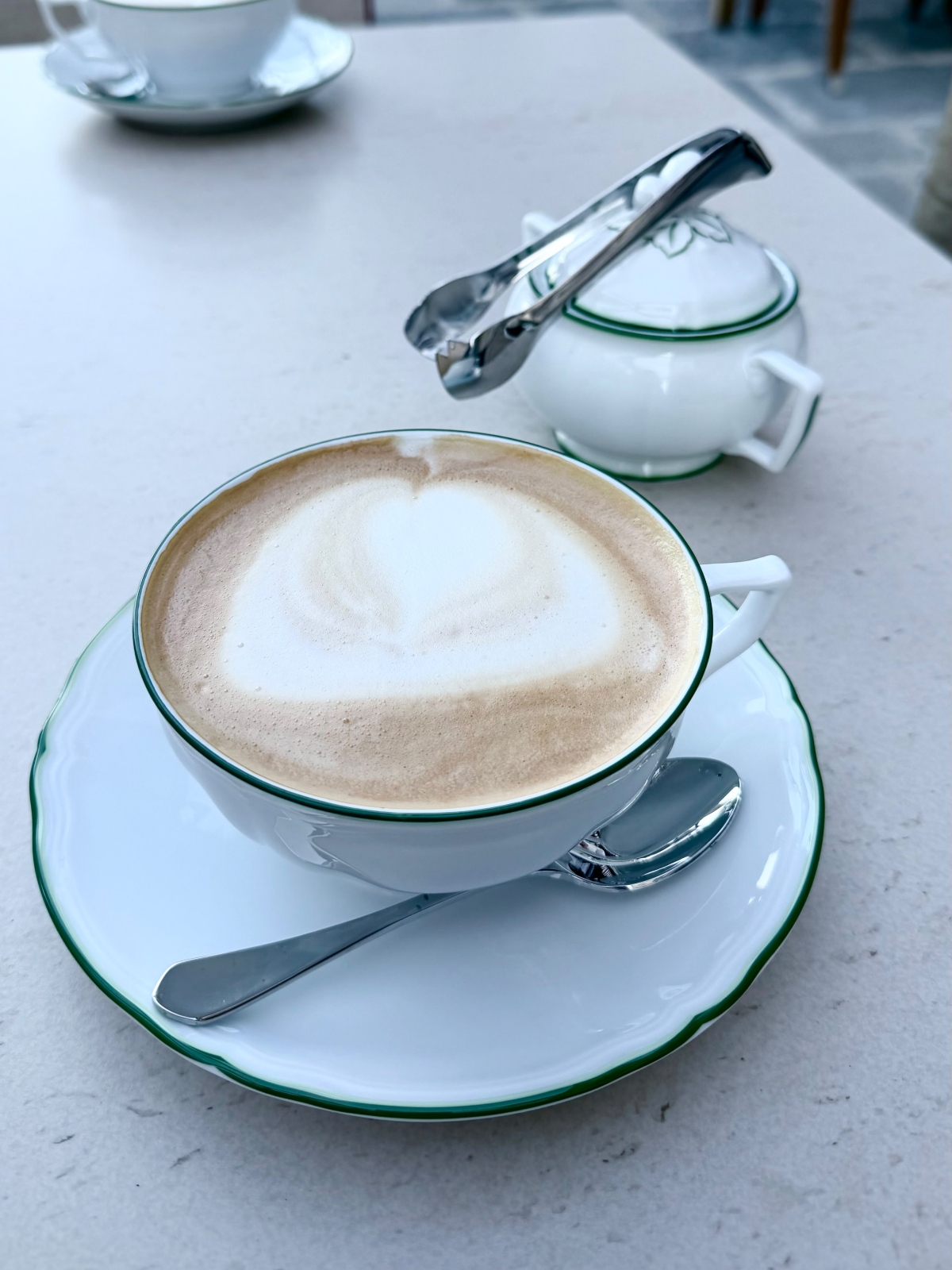 A cappuccino with heart-shaped latte art sits in a wide white porcelain cup with a thin green rim, resting on a matching saucer with a silver spoon. Behind it on the pale stone table, a lidded green-trimmed sugar bowl holds silver sugar tongs across its top, with a second cup and saucer in the background.