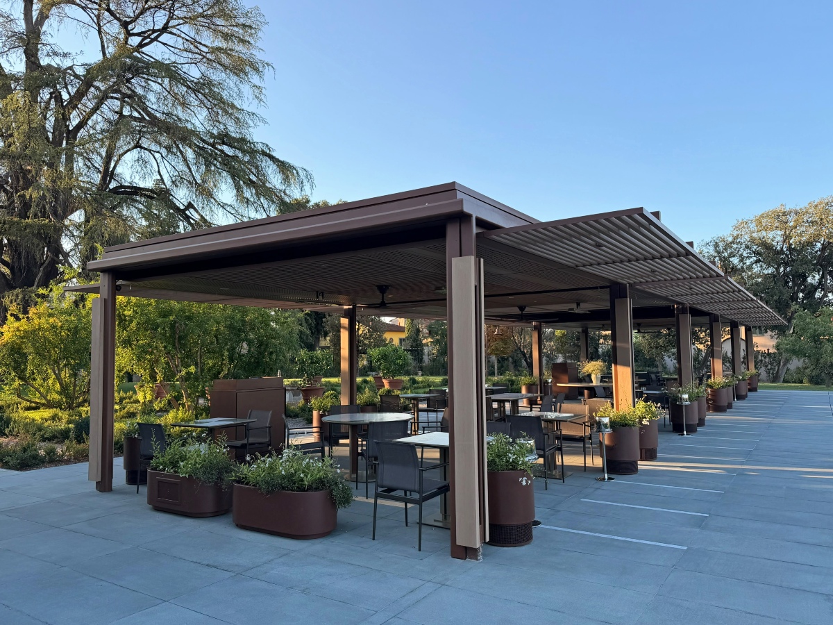 A modern outdoor dining terrace sits beneath a long bronze-toned pergola with a louvered roof, furnished with small tables, dark chairs, and large planters filled with greenery. The paved terrace is surrounded by lush gardens and mature trees, with soft golden light under a clear blue sky.