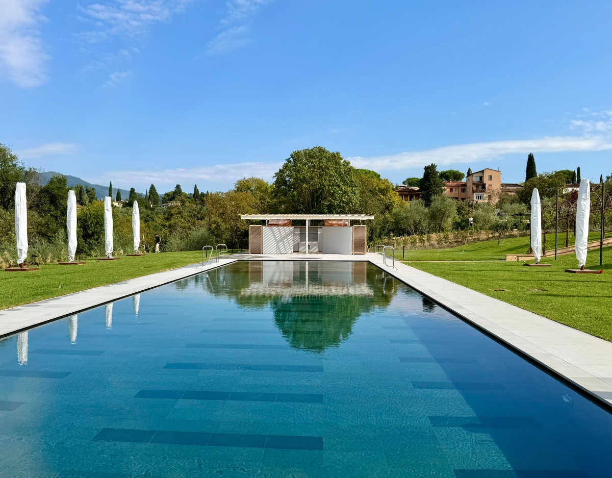 A long rectangular swimming pool with dark-tiled lane markings stretches toward a white pool house, its still blue water reflecting the trees and sky beyond. Manicured lawns with closed white umbrellas line both sides, set against a backdrop of cypress trees, terracotta-roofed buildings, and distant hills under a clear blue sky.