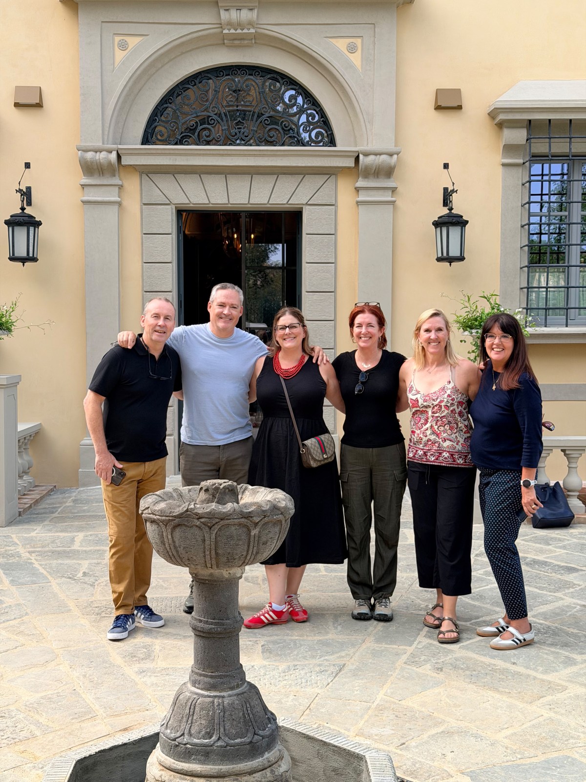 Six smiling adults stand arm in arm on a stone-paved courtyard in front of a pale yellow villa with a tall arched doorway crowned by an ornate wrought-iron fanlight. Black lantern sconces flank the entrance, and a carved stone fountain rises in the foreground.