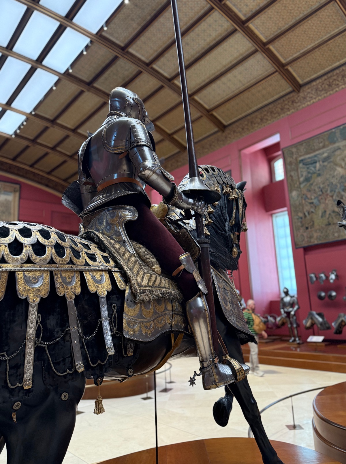 A full suit of steel armor mounted on a model horse holds an upright lance, with the horse draped in black velvet and silver-and-gold scalloped plates, tassels, and an embroidered saddle. The figure stands in a long gallery of the Royal Armoury in Madrid, with red walls, a vaulted ceiling with skylights, a large tapestry, and more armor and helmets displayed in the background.