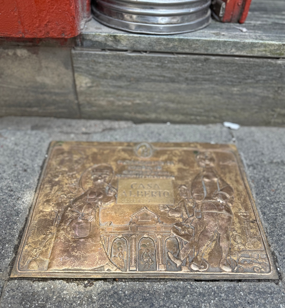 A bronze plaque set into a stone sidewalk reads "Casa Alberto, desde 1827," surrounded by raised relief figures in period dress and an arched building facade. The plaque sits in front of a stone step and a red-painted doorway, with a metal container resting on the step above.