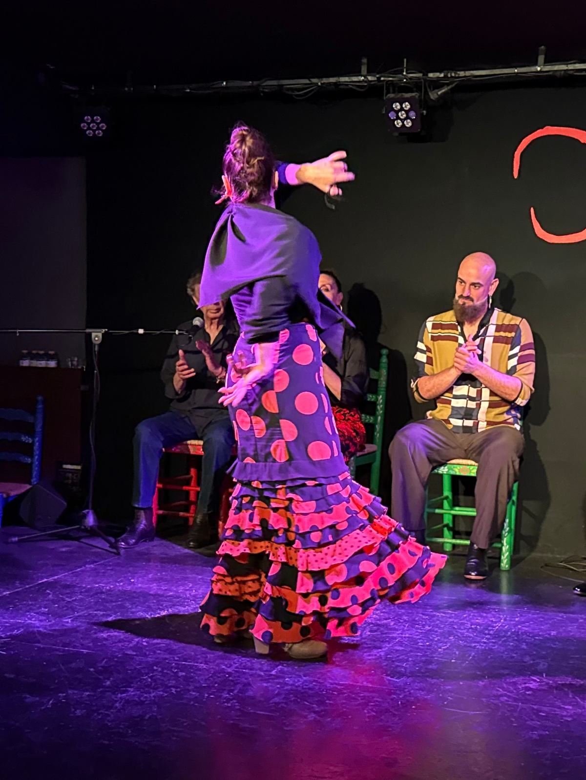 A flamenco dancer, seen from behind, twirls on a stage lit in purple, wearing a shawl and a ruffled polka-dot skirt in pink, orange, and black. Behind her, performers seated on painted wooden chairs clap along against a dark backdrop.