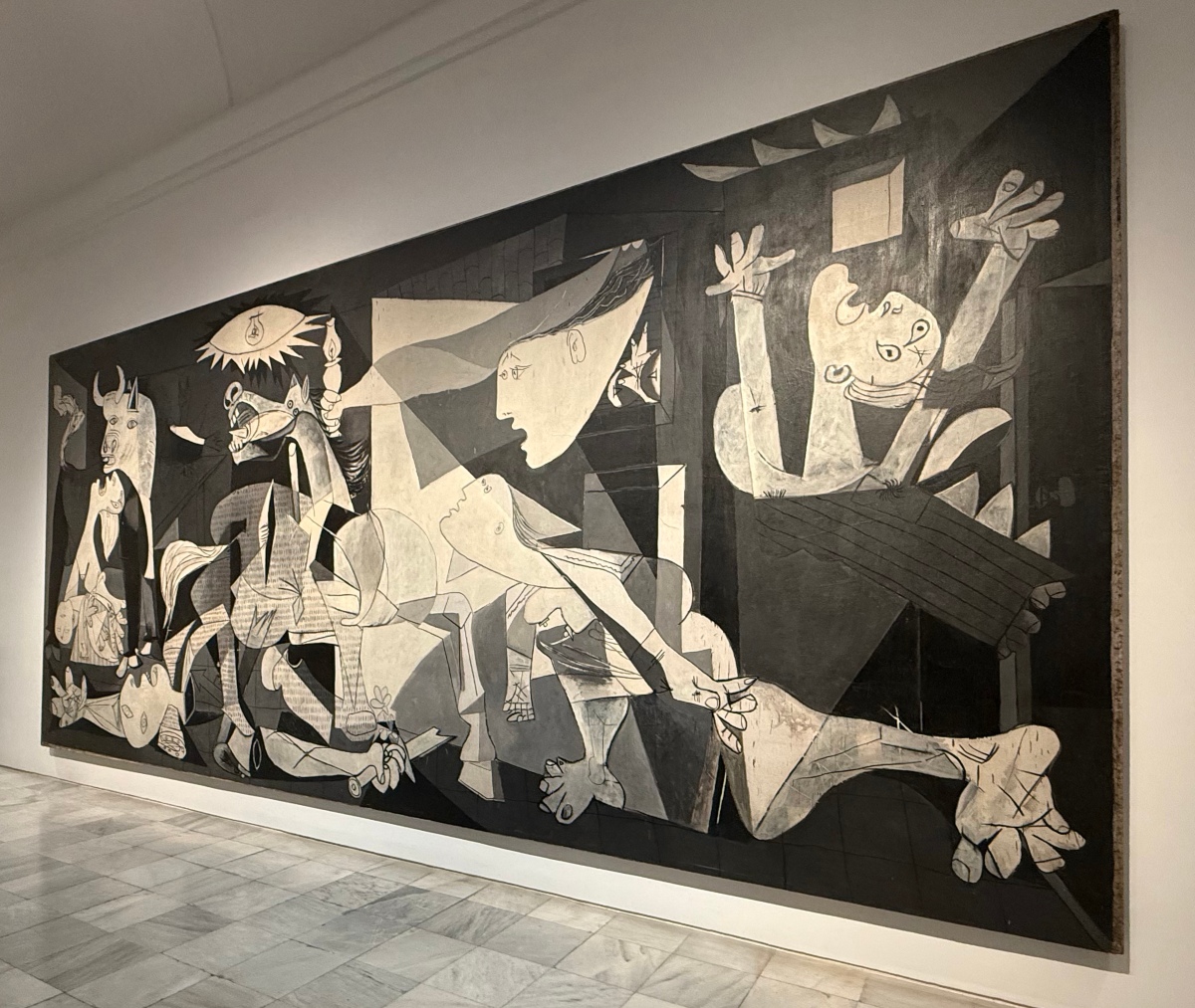 Pablo Picasso's *Guernica* fills a long white gallery wall at Madrid's Reina Sofía Museum above a gray marble floor. The large black, white, and gray painting shows fragmented, distorted figures, including a bull, an anguished horse, a woman holding a lamp, a figure with arms raised, and a fallen soldier, beneath a light shaped like an eye.