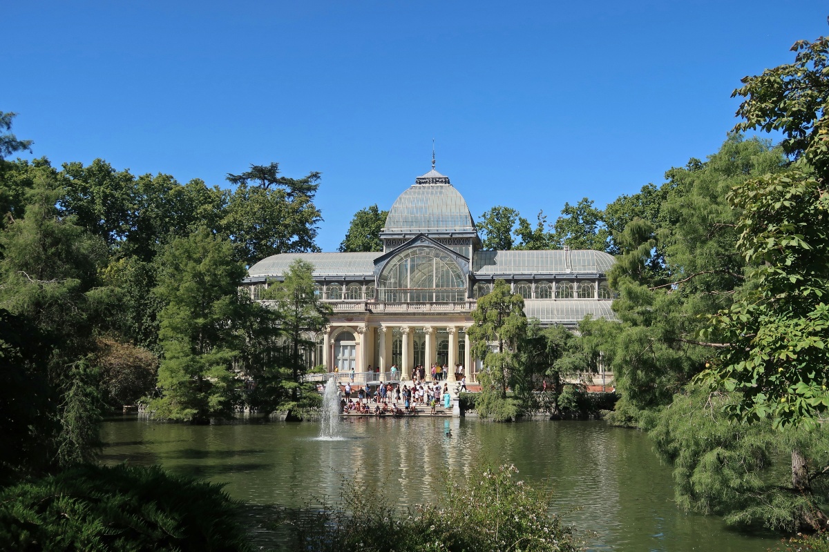 The Crystal Palace in Madrid's Retiro Park, a glass-and-iron pavilion with a domed roof and a columned entrance, stands behind a green pond with a fountain spraying in the center. Visitors gather on the steps leading down to the water, and tall trees frame the scene under a clear blue sky.