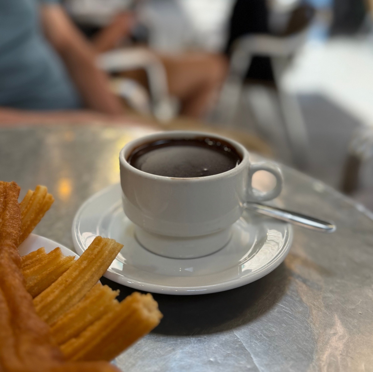A white cup of thick hot chocolate sits on a saucer with a spoon on a metal café table, beside a plate of golden fried churros in the foreground. In the blurred background, people sit at tables in the café.