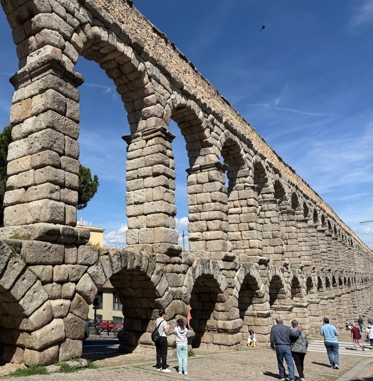 The Roman Aqueduct of Segovia stretches into the distance under a bright blue sky, its two tiers of arches built from large, stacked granite blocks. Visitors walk and stand along the cobblestone plaza at its base, dwarfed by the towering stone pillars.