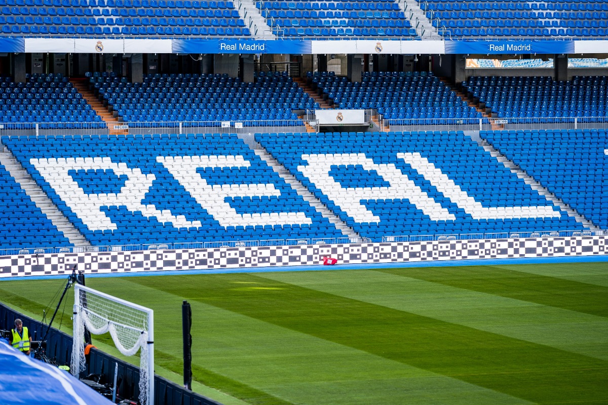 Rows of empty blue stadium seats at Real Madrid's Santiago Bernabéu Stadium are arranged so white seats spell out "REAL" in large letters, with Real Madrid banners lining the upper tier. In the foreground, a striped green soccer pitch stretches toward a goal net, with a worker in a high-visibility vest standing near the sideline.