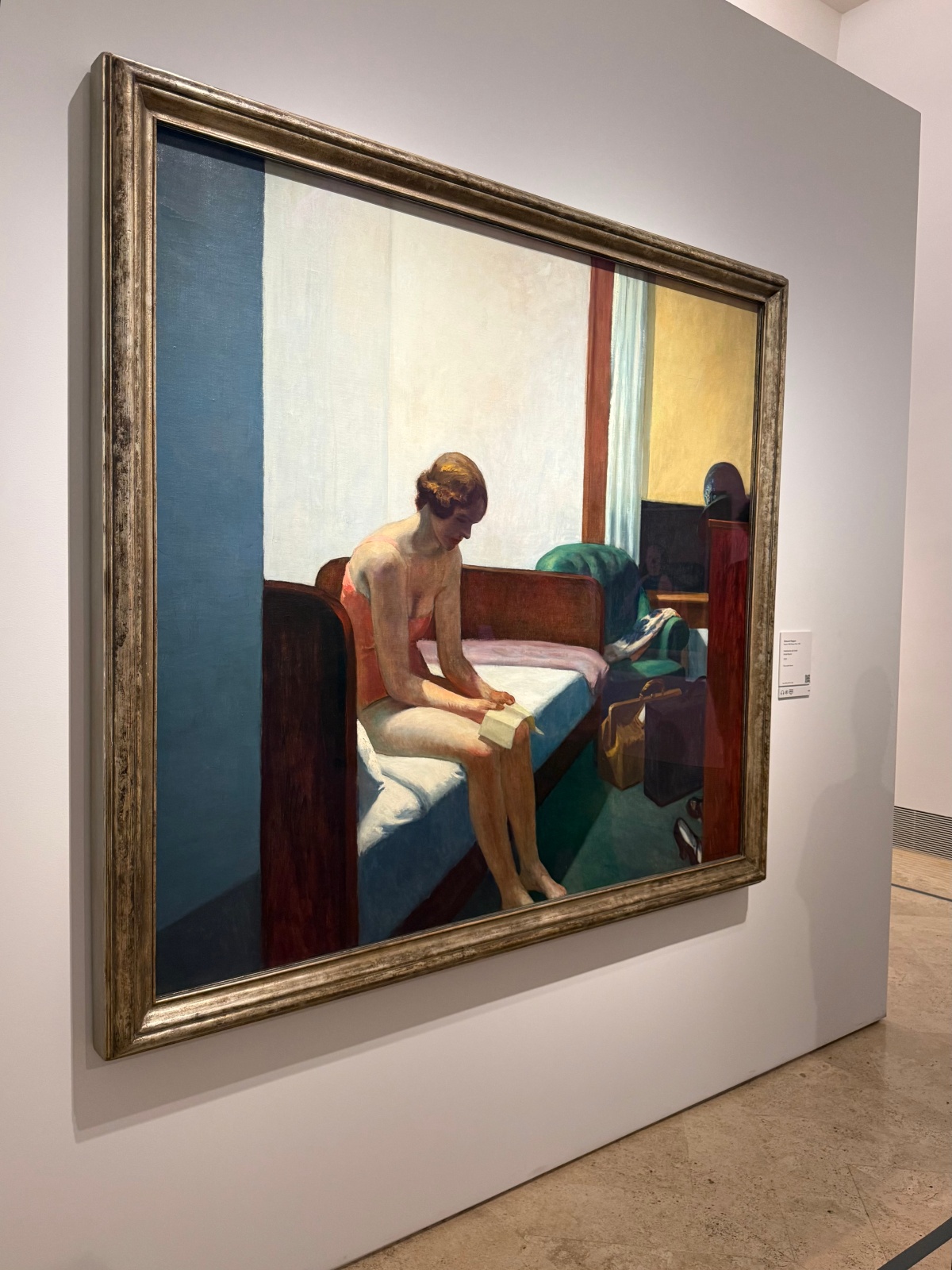 Edward Hopper's painting *Hotel Room* hangs in a wide silver frame on a white gallery wall at Madrid's Thyssen-Bornemisza Museum, with a small label beside it. The painting shows a woman in a pink slip sitting on the edge of a bed, reading a slip of paper, with a green armchair, luggage, and shoes nearby in the sparsely furnished room.