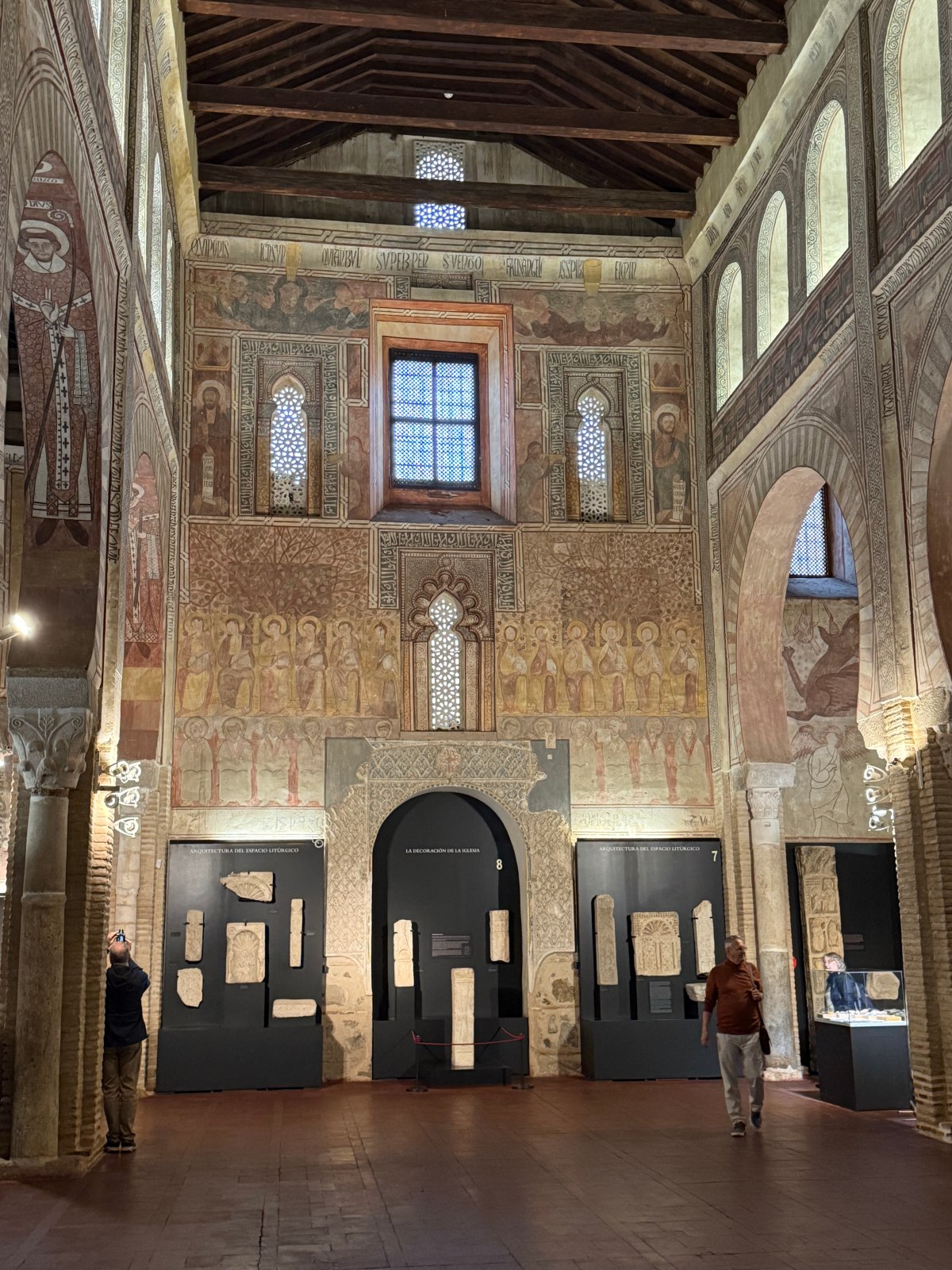 Here's the updated alt text:The interior of the Church of San Román in Toledo features walls covered in faded medieval frescoes of saints and robed figures, along with Moorish-style horseshoe arches, latticed windows, and inscriptions beneath a dark timber ceiling. Stone carvings are displayed on dark museum panels along the brick floor, where a few visitors walk and take photos.