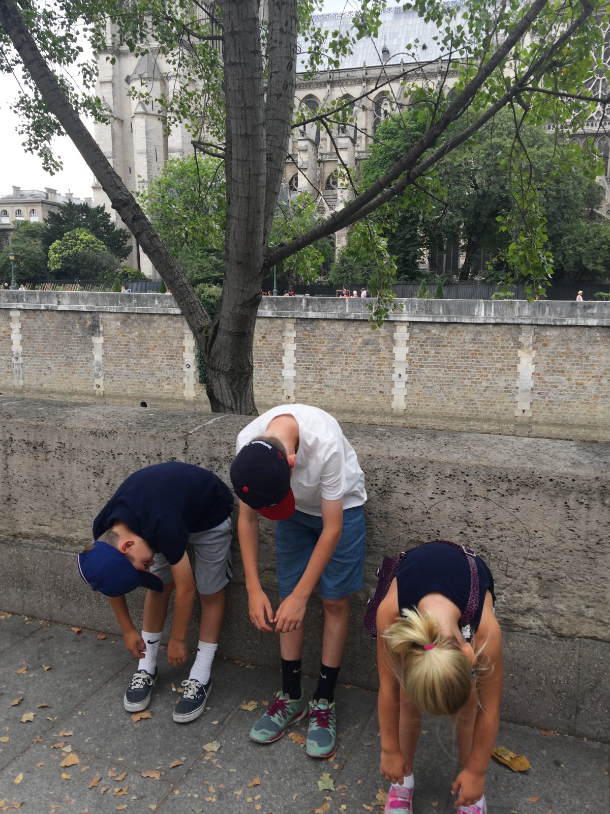 Three children in shorts and sneakers bend over at the waist with their heads down beside a low stone wall along a riverside walkway, two of the boys wearing navy baseball caps. Behind them, a tree arches over a brick embankment, with the Gothic towers and stone facade of Notre-Dame Cathedral in Paris rising in the background.