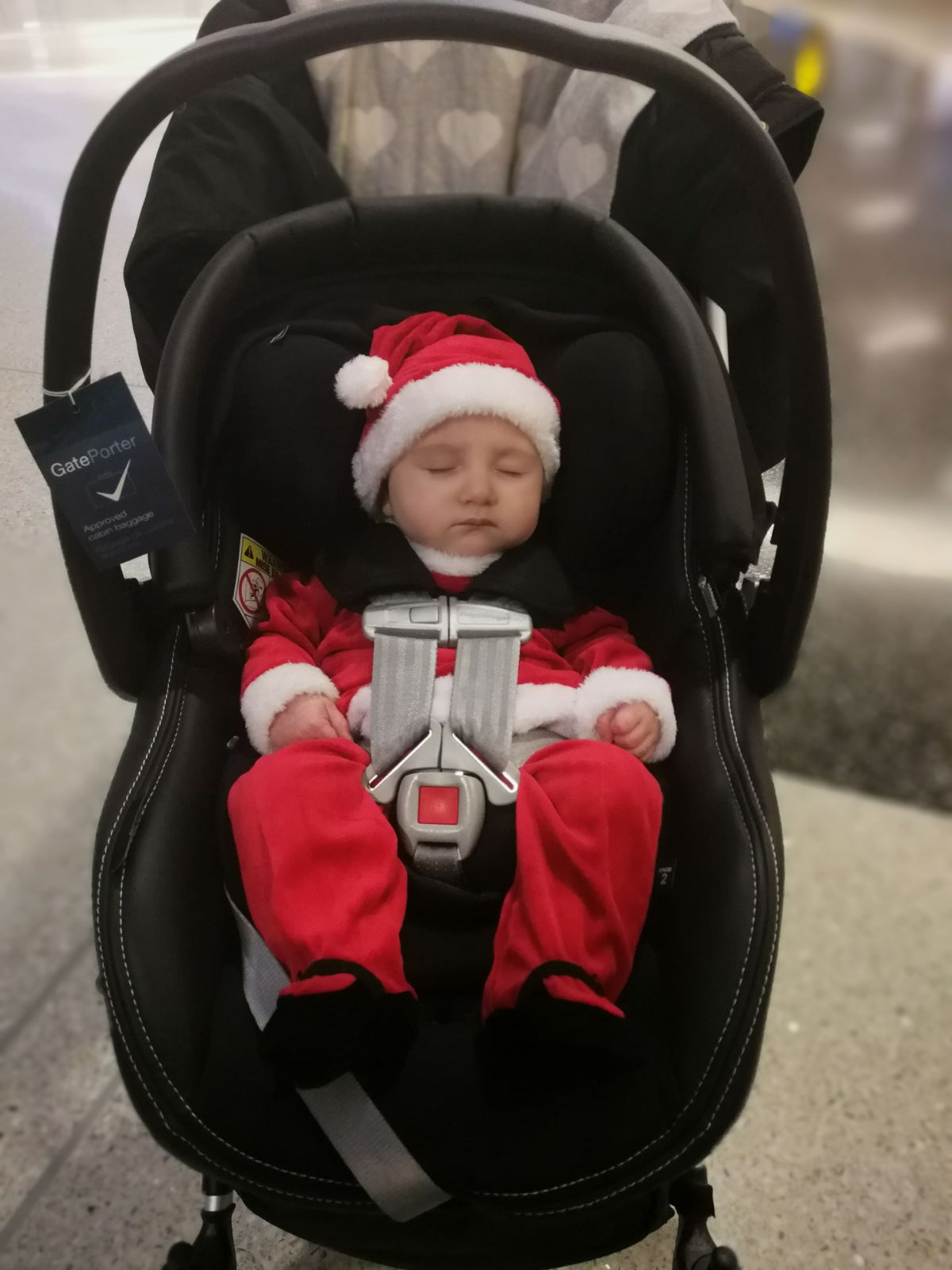 A sleeping baby in a red-and-white Santa suit and matching hat is buckled into a black infant car seat mounted on a stroller. A "GatePorter – Approved cabin baggage" tag hangs from the car seat handle, and the setting appears to be an airport terminal floor.